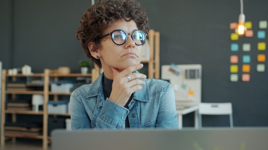 Person in glasses thinking deliberately at a work desk while preparing a job search with a laptop nearby