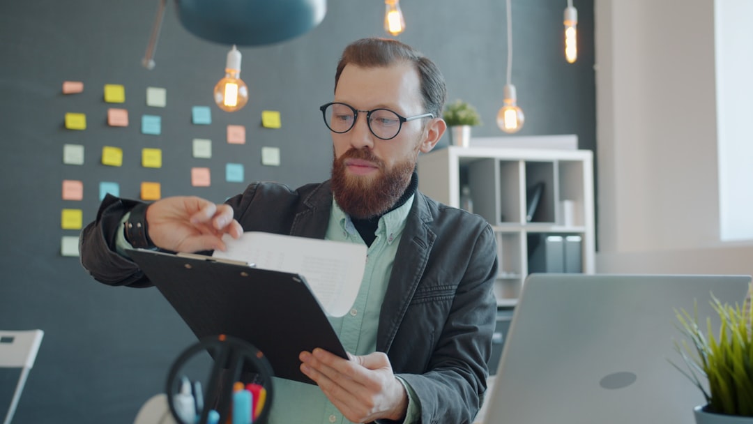 A job candidate in glasses sits at his desk and reads carefully through the pages of an offer document on a clipboard before signing it.
