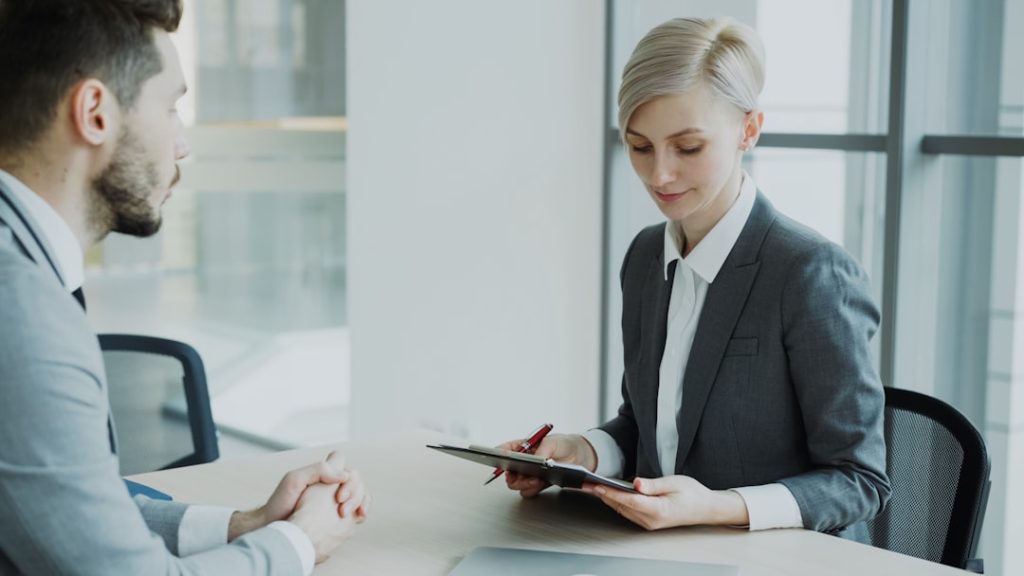 A hiring manager in a suit reviews a candidate document across a table while the candidate sits waiting with hands clasped, the moment a resume gap comes up in an interview.