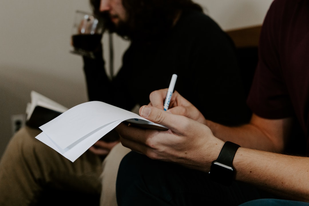 An interviewer writes notes on a sheet of paper while the person they are interviewing sits across from them, out of focus in the background.