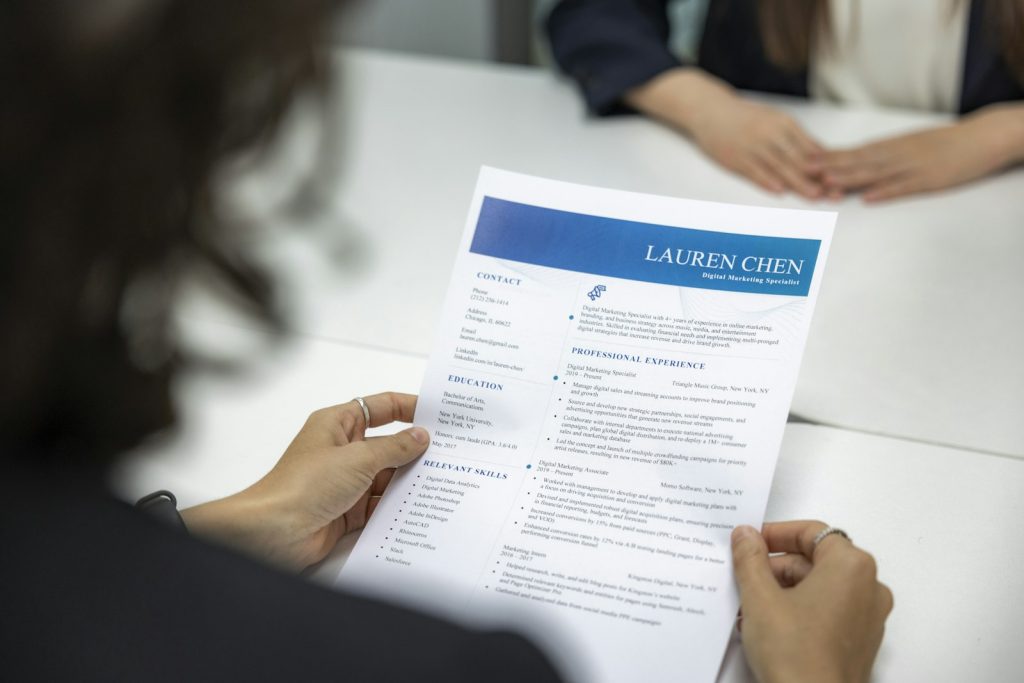 A woman sits at a table reading a printed resume closely, reviewing it line by line