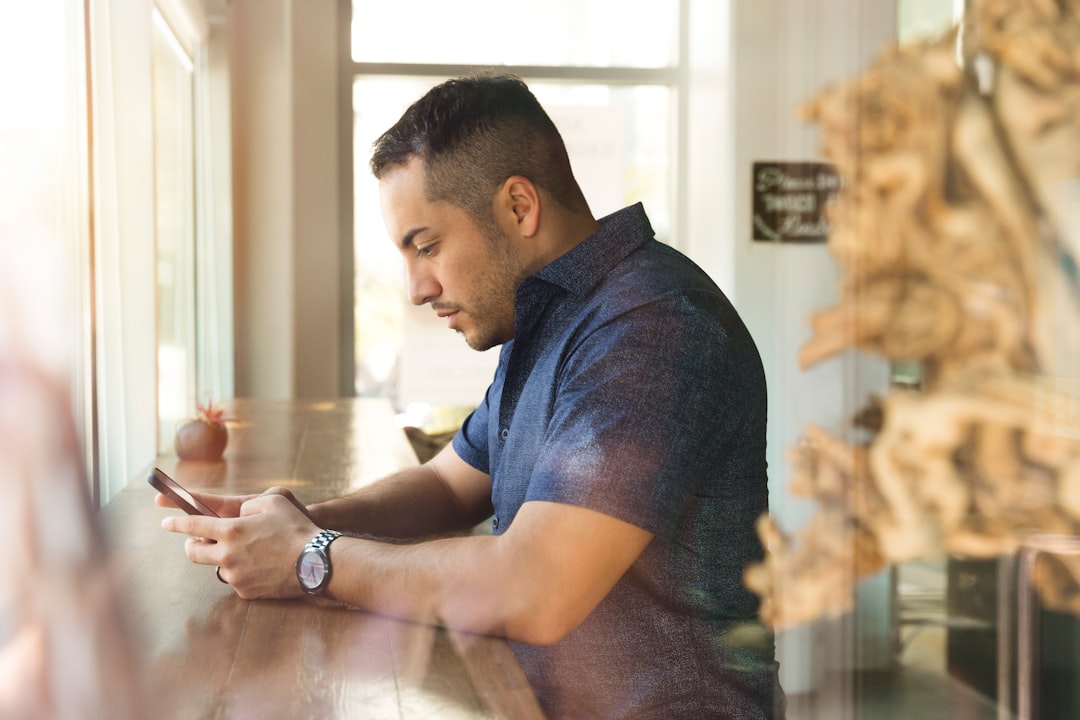 A professional in a dark button-up shirt sits at a window counter, focused on composing a follow-up message on his phone after a recruiter went quiet.