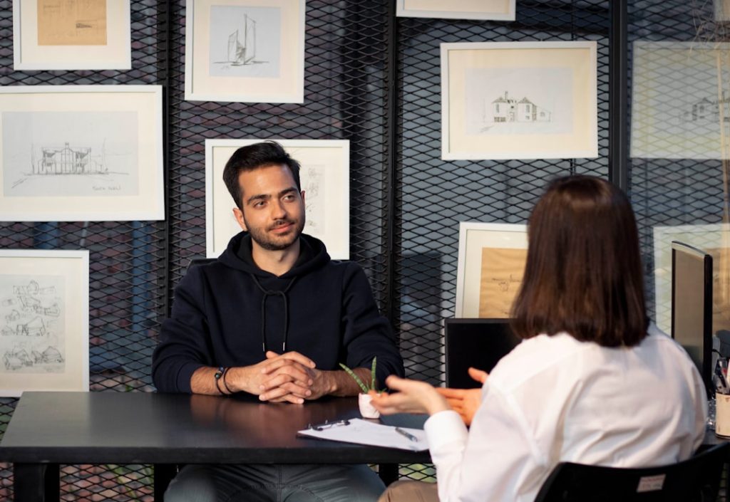 A job candidate sits across a table from an interviewer holding a clipboard, pausing to think before answering a question.