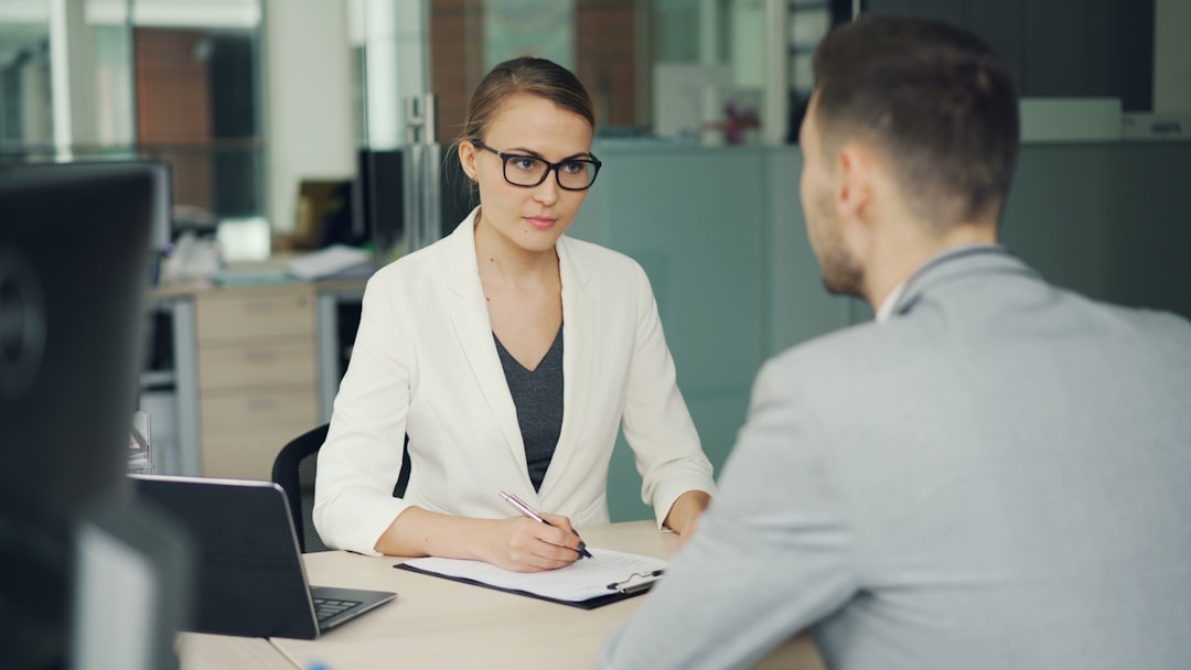 A job interviewer in glasses takes notes on a clipboard while listening to a candidate seated across the desk from her.