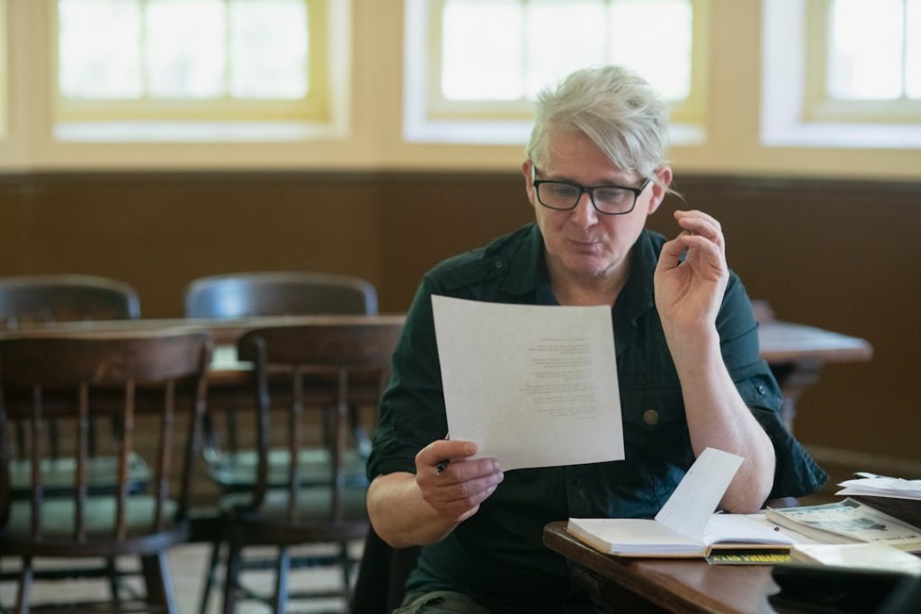 A grey-haired professional sits at a wooden table reading a single-page letter, a pen in one hand and an open notebook beside him, weighing what it says.