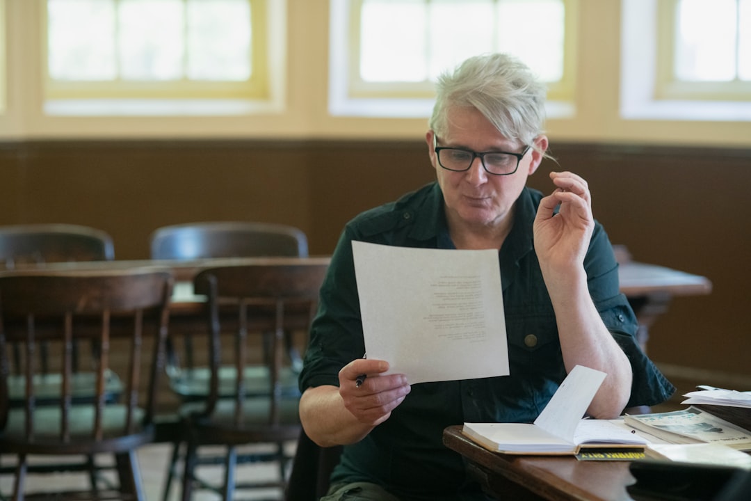 A grey-haired professional sits at a wooden table reading a single-page letter, a pen in one hand and an open notebook beside him, weighing what it says.