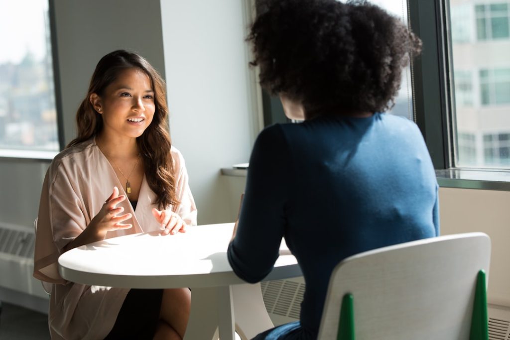Two women seated at a small round table by an office window, one gesturing with her hands as she answers a question while the other listens.