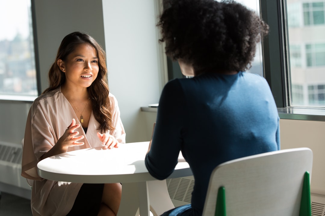 Two women seated at a small round table by an office window, one gesturing with her hands as she answers a question while the other listens.