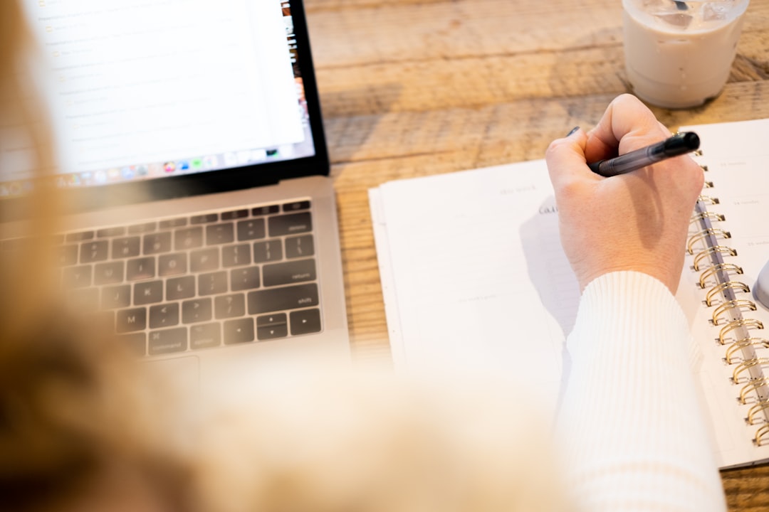 Over-the-shoulder view of a person handwriting notes in a spiral planner beside an open laptop on a wooden table.