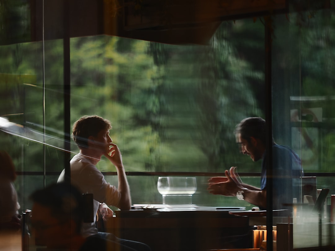 Two people seen through a window in an intense conversation across a table, one gesturing mid-explanation while the other listens