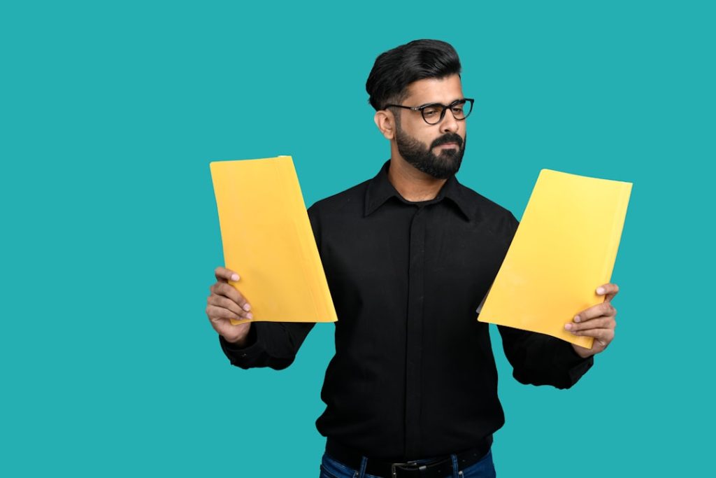 A bearded man in a black shirt holds a yellow folder in each hand against a teal background, looking at one as he weighs the two against each other.