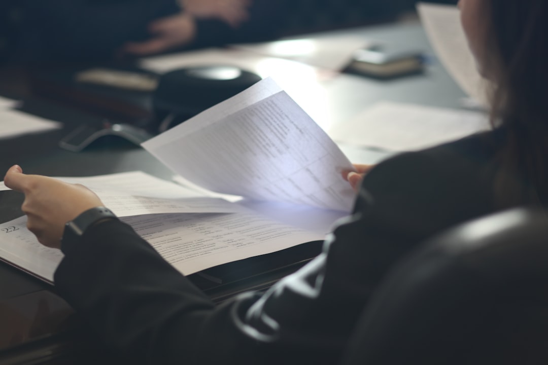 A professional in a dark suit turns the pages of a printed document at a meeting table, with a conference phone and more paperwork spread nearby.