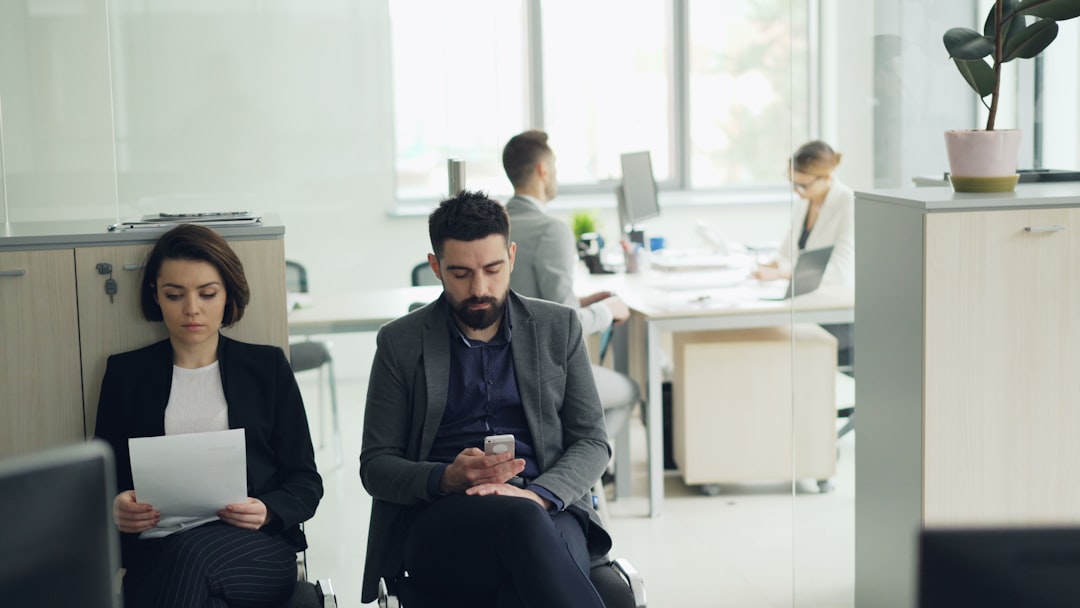 Two job candidates in business attire wait on chairs in an open-plan office, one holding printed pages and one checking a phone, while two employees work at desks behind them.
