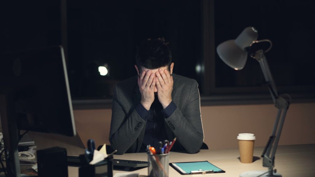 A man in a suit sits alone at a desk late at night with his head in his hands, a computer monitor and desk lamp beside him.
