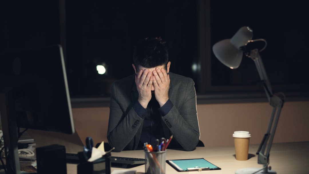 A man in a suit sits alone at a desk late at night with his head in his hands, a computer monitor and desk lamp beside him.