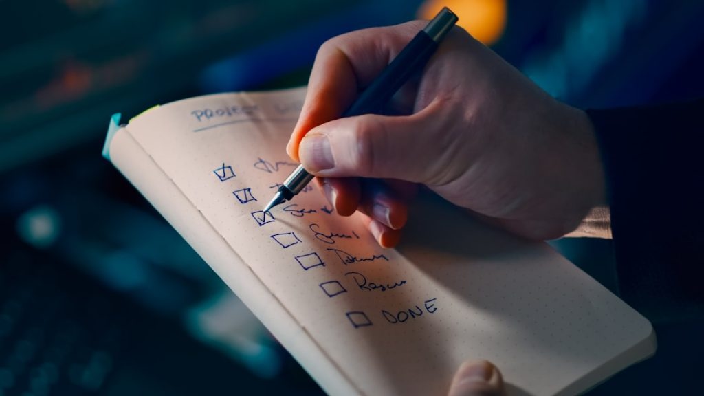 A hand ticks off a handwritten checklist of steps in a notebook, working down the page toward DONE.