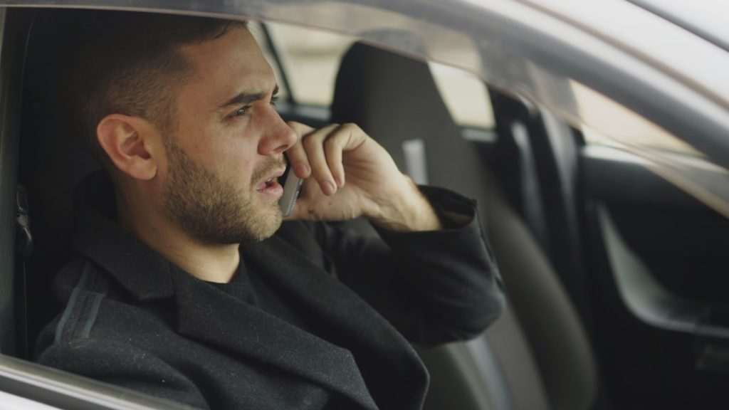 A man in a dark overcoat sits in a parked car taking a phone call, the way employed candidates handle interview calls during the workday.