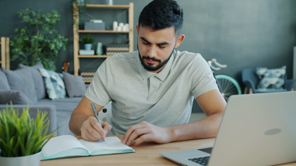 A man in a grey polo shirt sits at a light wood table in a home living room, writing in an open notebook with a pen while an open laptop sits beside him.