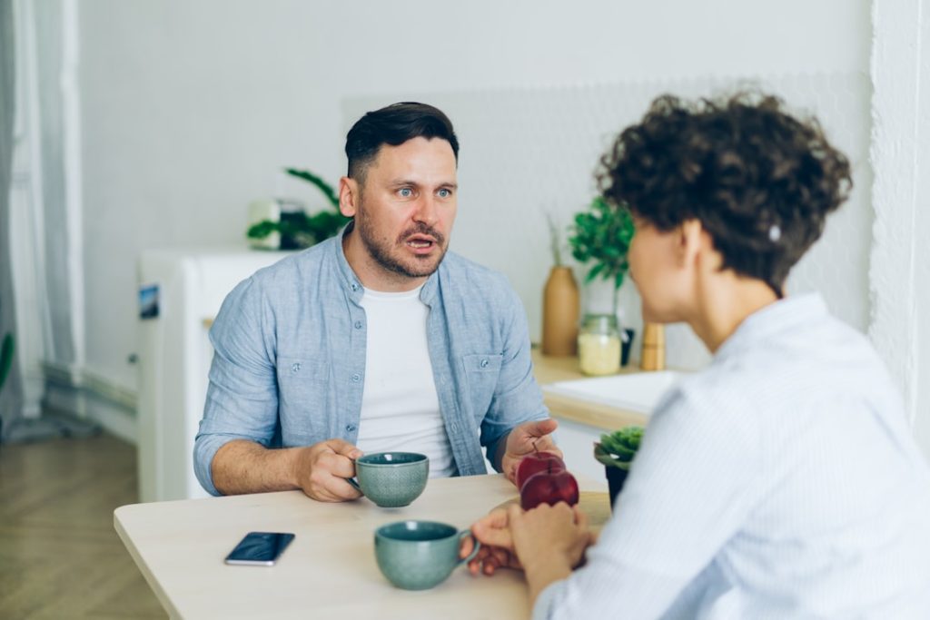 A man sits at a table mid-sentence, mouth open and hand gesturing as he explains something, while a woman across from him listens in silence.