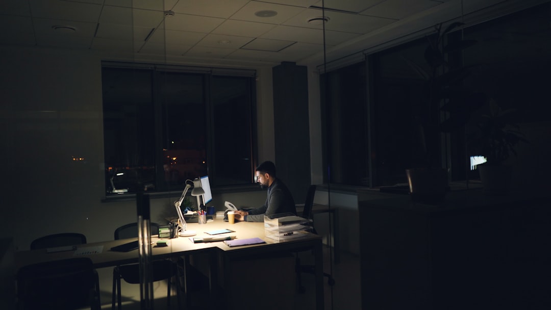 A man works alone at a lit desk in a dark, empty open-plan office at night, surrounded by unoccupied chairs and desks.