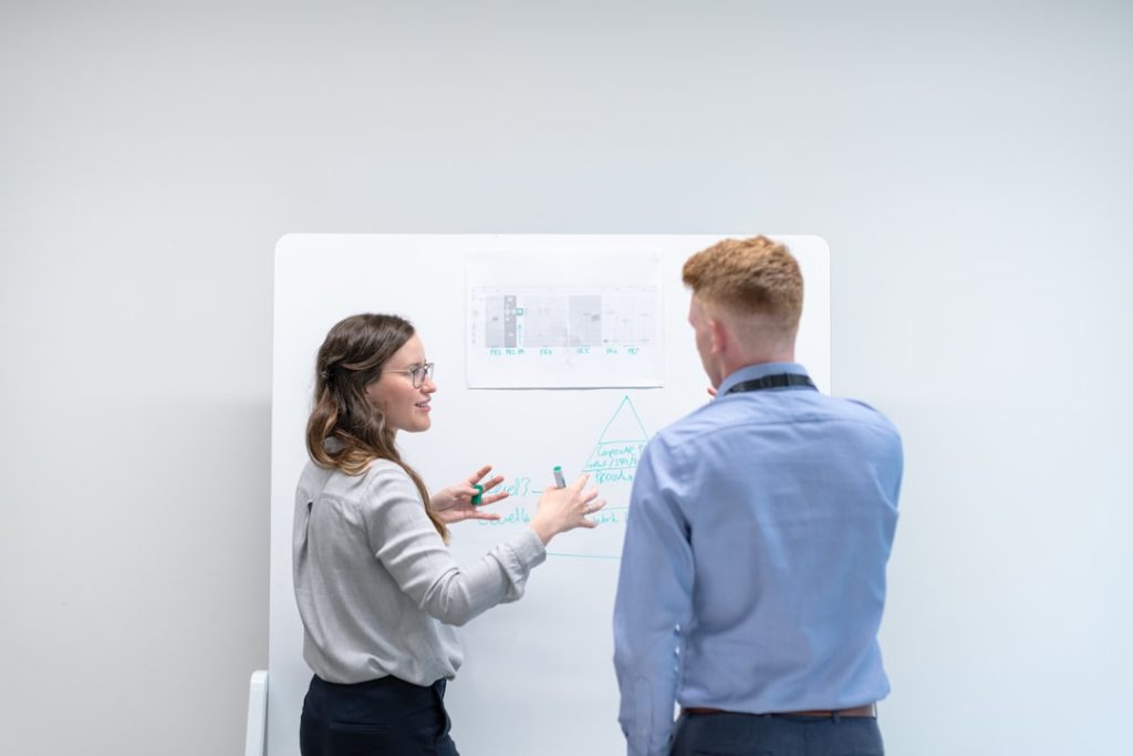A candidate explains her reasoning at a whiteboard covered in technical diagrams while an interviewer listens.