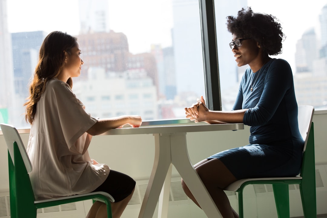 Two professionals sitting across a small table by a window, facing each other mid-conversation during a job interview.