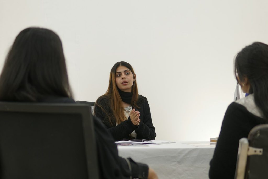 A candidate sits across a table from a two-person interview panel, hands clasped, mid-sentence as she explains her work
