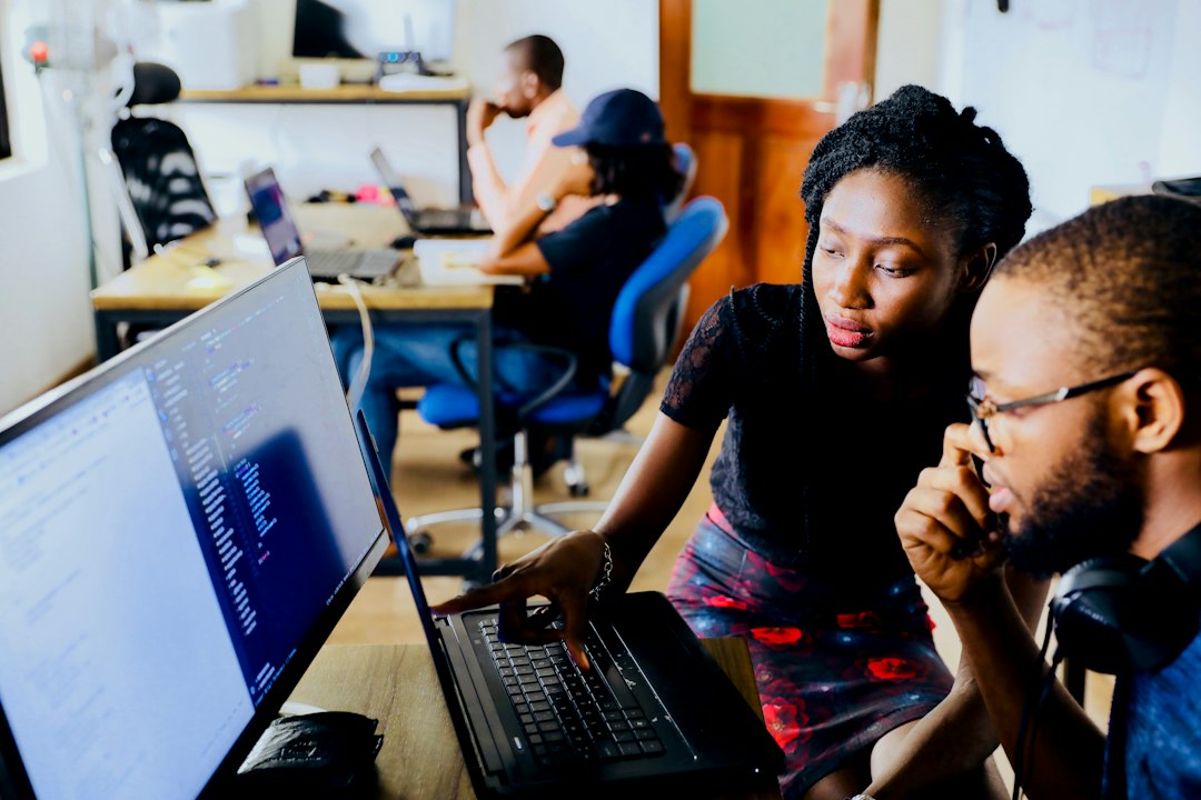Two software engineers review code together at a desk, one pointing at the laptop while the other listens and weighs the call, with code on a large monitor beside them