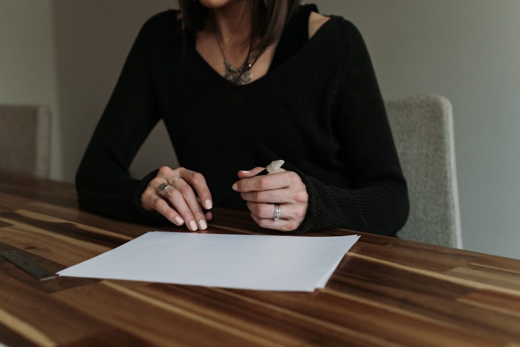 A woman in a black sweater sits at a wooden table holding a pen over a blank sheet of paper, pausing before she writes
