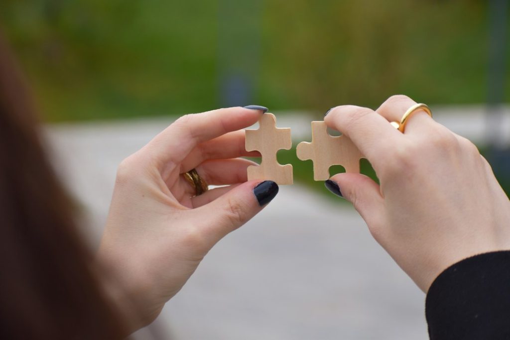 Two hands holding two wooden jigsaw puzzle pieces up to each other, about to fit together, against a blurred green outdoor background