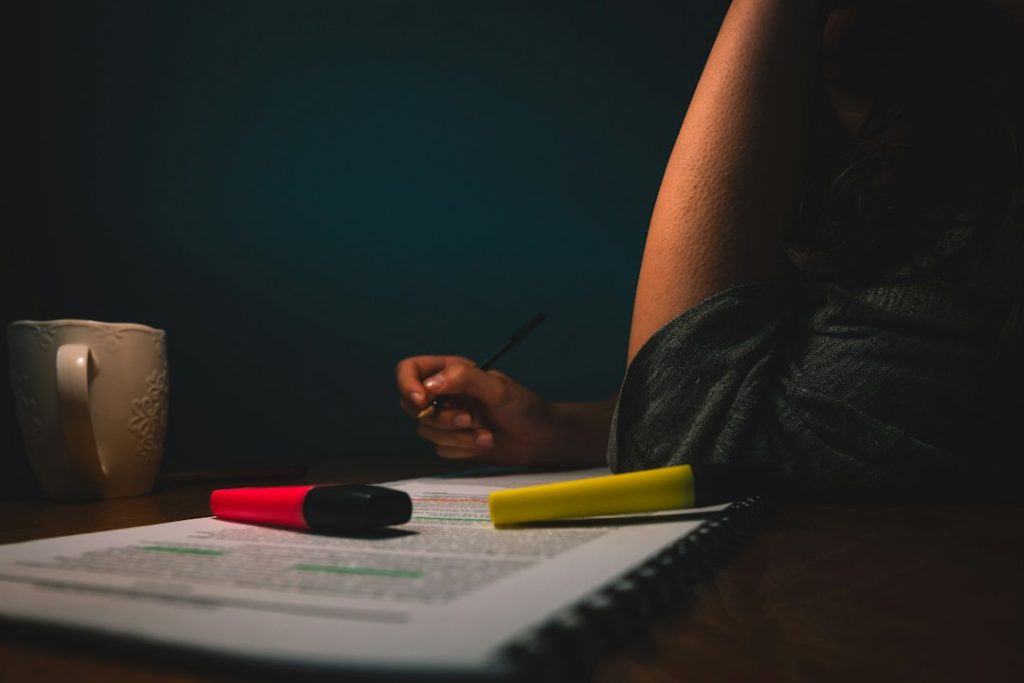 A person sits at a dark desk with a pen in hand, working through a printed document marked up with red and yellow highlighters, a coffee mug beside them - preparing before an interview.
