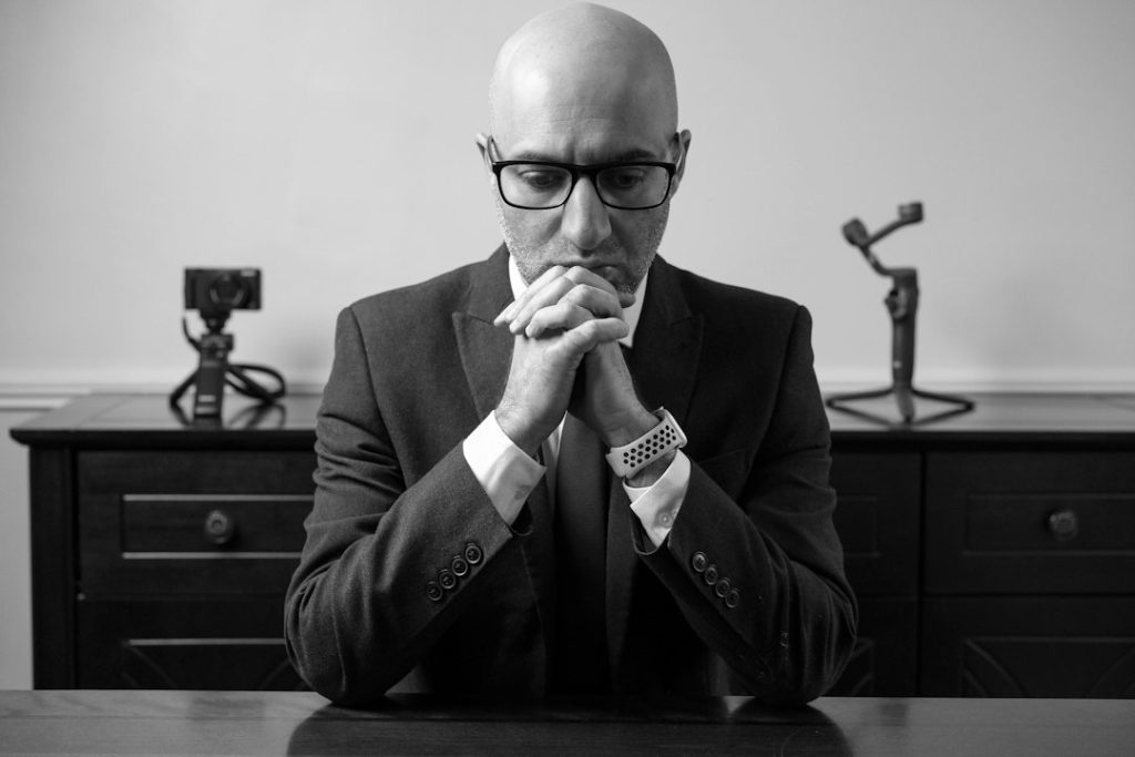 Man in a dark suit and glasses sitting at a table with his hands clasped in front of his mouth, tense before a job interview