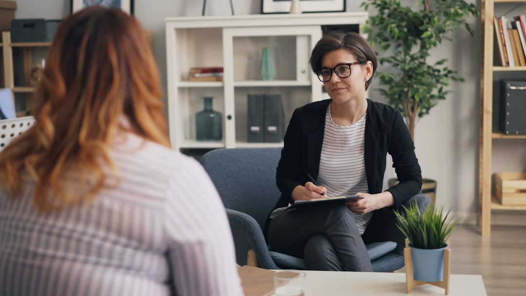 Interviewer in glasses listening closely and taking notes on a clipboard while a candidate, seen from behind, answers across a small table