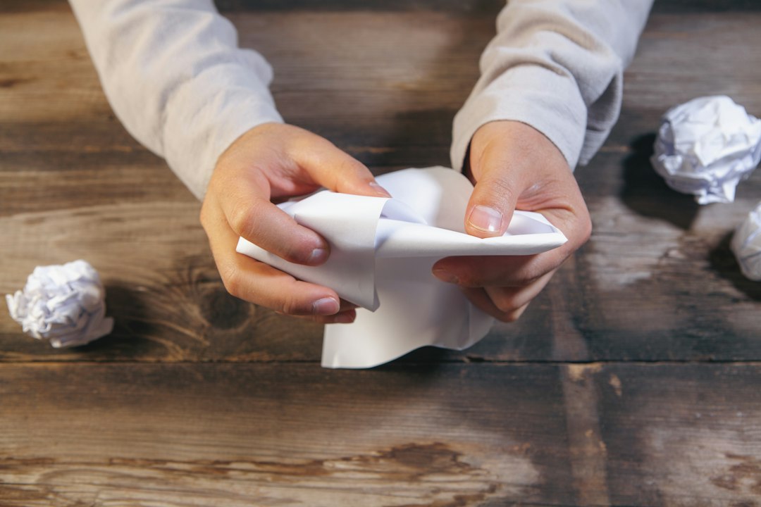 Two hands crumpling a sheet of paper over a wooden desk, with other crumpled paper balls nearby - discarding a job search plan that stopped working