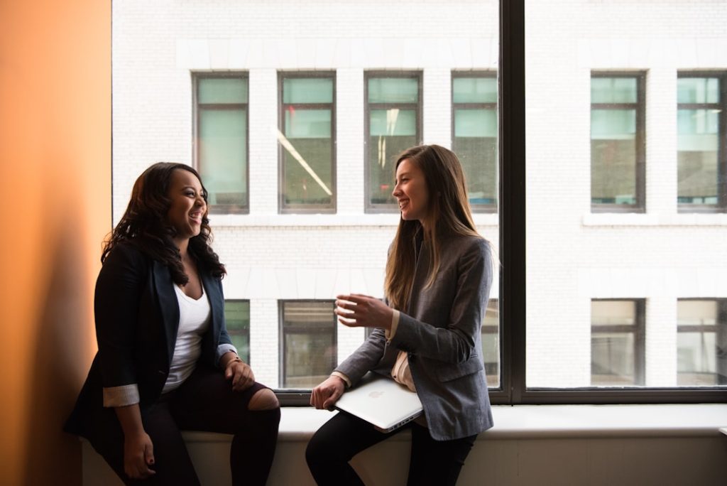 Two professionals sitting on an office windowsill, laughing mid-conversation, one holding a closed laptop - an easy, genuine networking chat