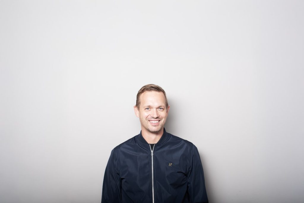 Smiling man in a dark jacket in front of a plain light gray wall, lit evenly and looking straight at the camera - the kind of simple, direct LinkedIn profile photo that earns the click