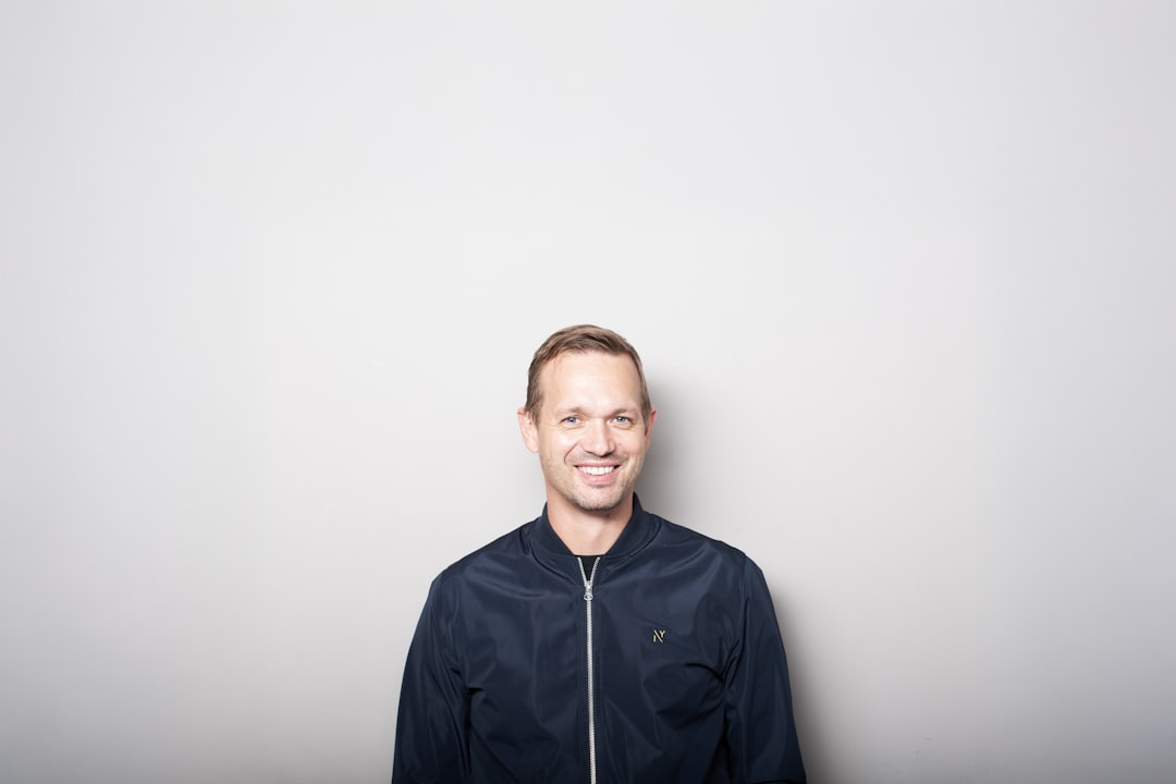 Smiling man in a dark jacket in front of a plain light gray wall, lit evenly and looking straight at the camera - the kind of simple, direct LinkedIn profile photo that earns the click