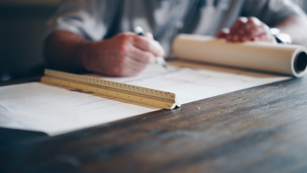 Person drafting a plan by hand on a roll of paper at a wooden desk, with a scale ruler in the foreground - building the one-page proof of work before an interview