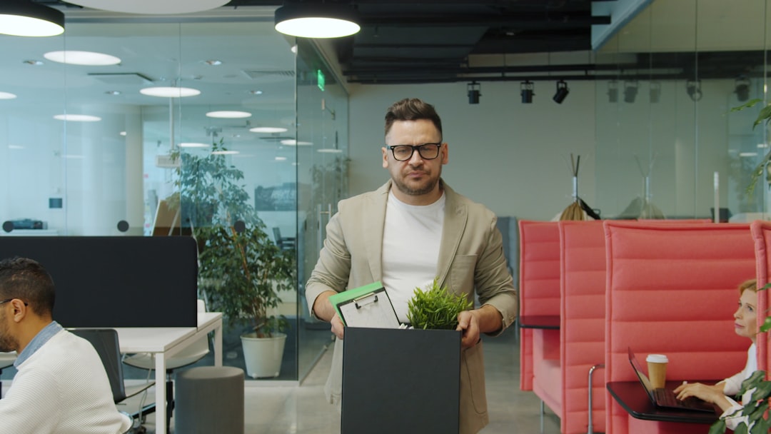 A man in glasses and a beige blazer carries a box of his desk belongings, a plant and a clipboard on top, through an open-plan office as coworkers keep working