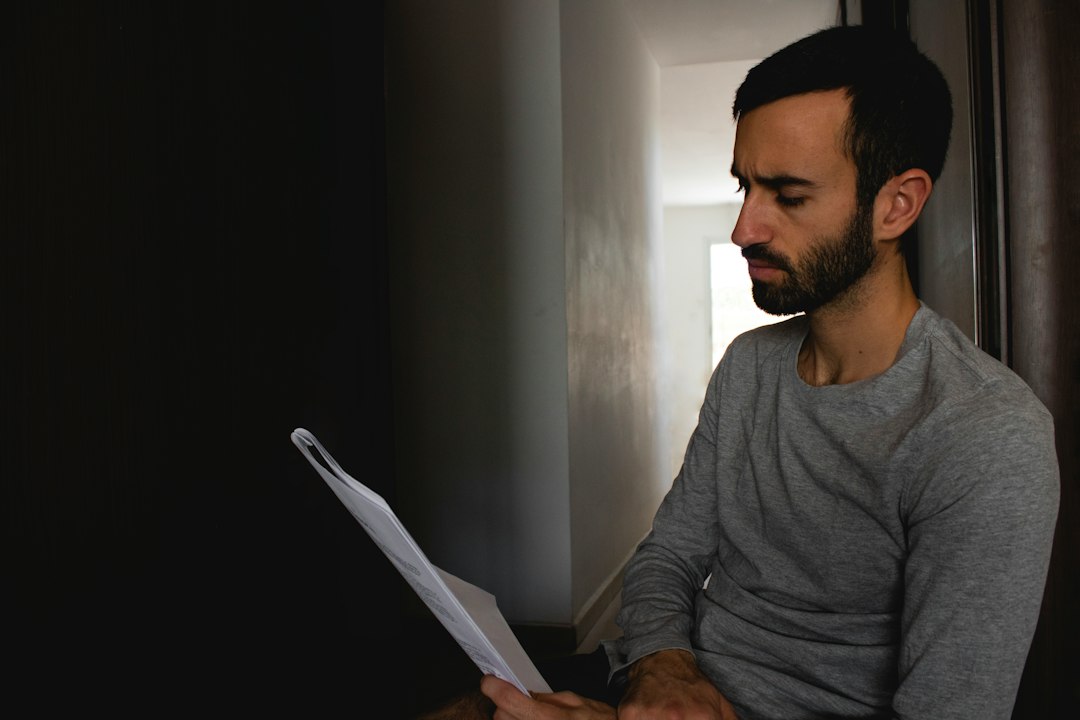 A man in a gray sweater sits by a doorway, reading a printed document closely with a serious expression.