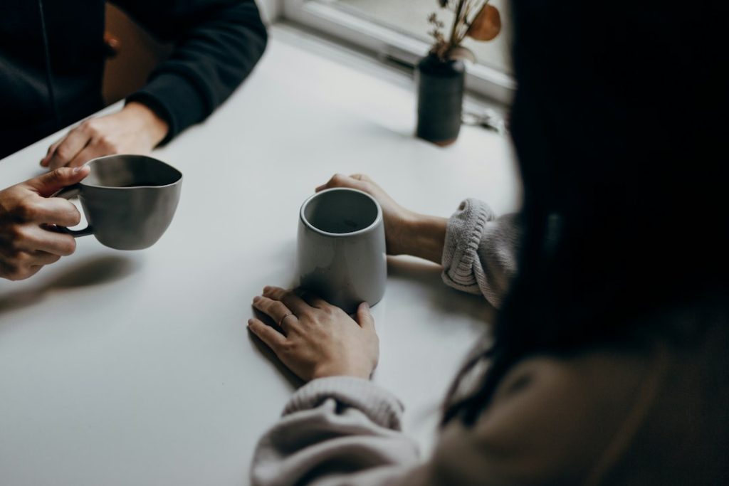 Two people sitting across a white table by a window, hands wrapped around grey coffee mugs, in a quiet one-on-one conversation