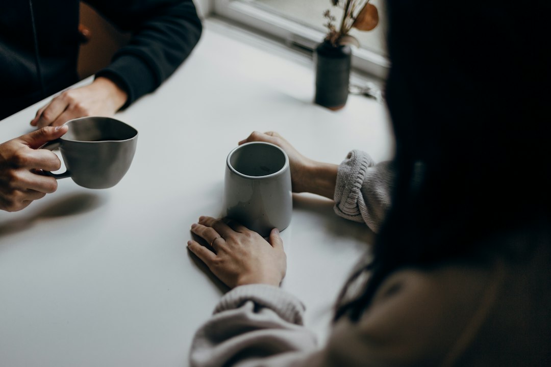 Two people sitting across a white table by a window, hands wrapped around grey coffee mugs, in a quiet one-on-one conversation