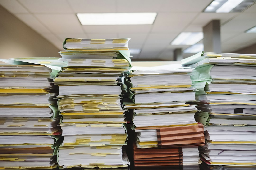 Tall, uneven stacks of paper files and folders piled on an office desk, representing the growing pile of job applications recruiters sort through for every hire