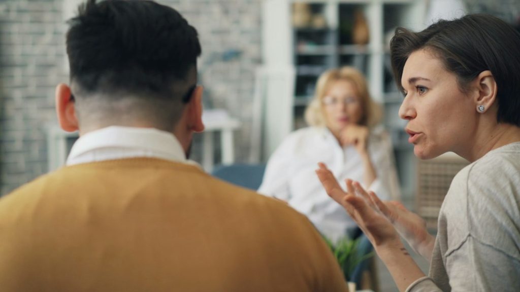 A woman gestures as she explains her point to a man seated with his back to the camera, while a third person watches in the background.