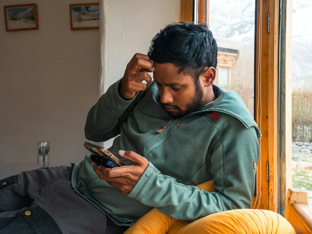 A man in a green fleece sits by a window, one hand to his forehead, frowning at the phone in his other hand