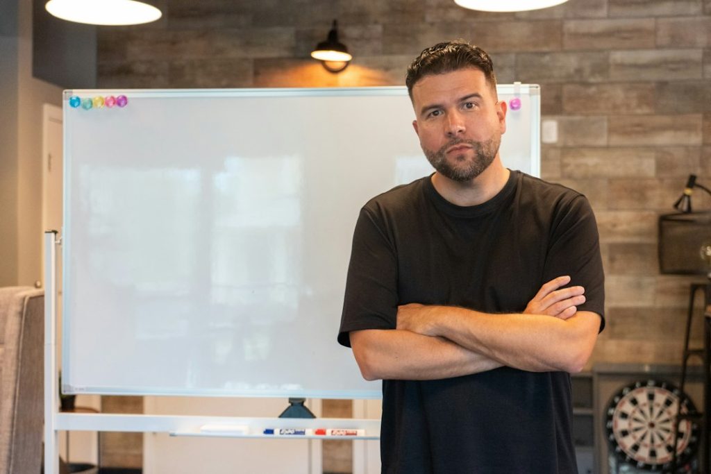 A man in a black t-shirt stands with arms crossed beside a blank whiteboard, looking at the camera.