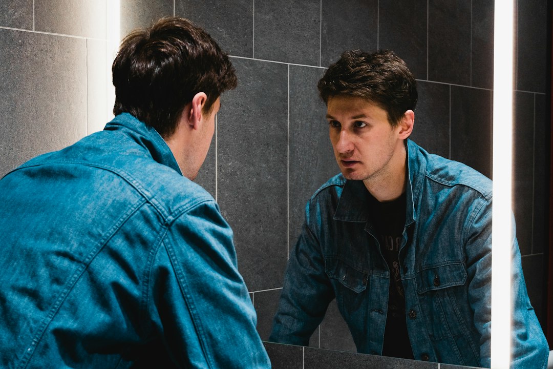 A man in a denim jacket studies his own reflection in a bathroom mirror with a serious, thoughtful expression