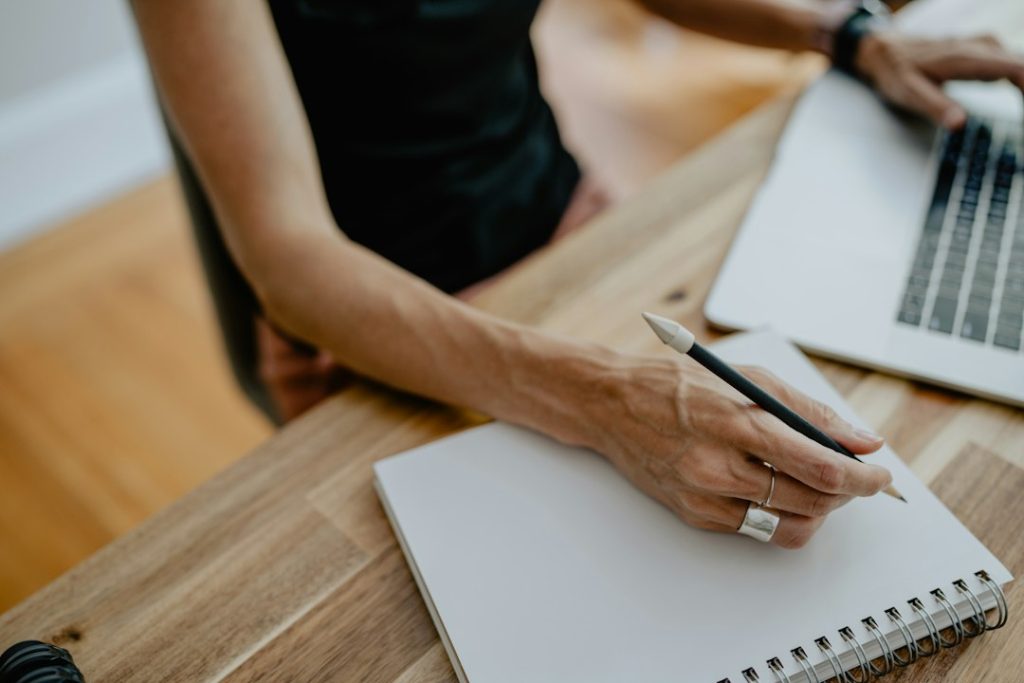 Hand holding a pencil over a blank spiral notebook on a wooden desk, a laptop open beside it