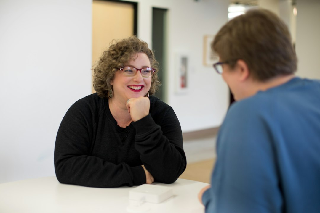 Two people talking across a white table, one smiling and listening closely with her chin resting on her fist