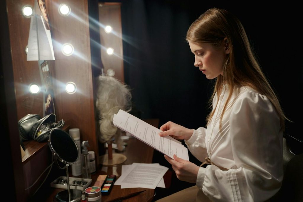 A woman in a white blouse studies a stack of printed pages at a dressing-room table lit by bulb lights, rehearsing before a performance.